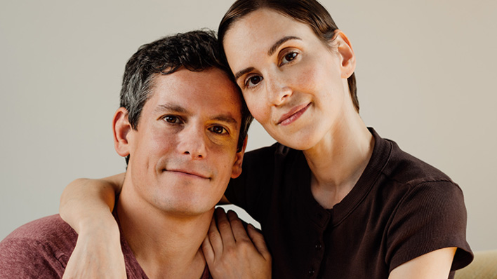 Two people posed closely together, one with an arm around the other's shoulder, facing the camera against a plain background.