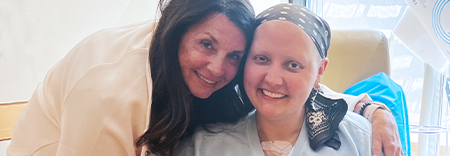 Two people smiling together in a medical setting, one wearing a headscarf and the other with long dark hair, seated near hospital equipment