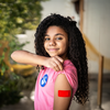 Young girl showing a red bandage on her arm after receiving a shot, smiling confidently.