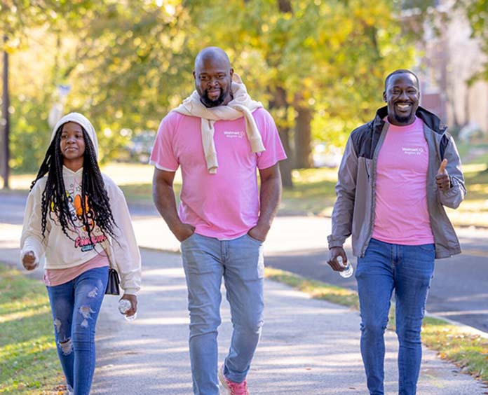 Three people walking together on a sunny day in a park, two wearing pink t-shirts and jeans, suggesting participation in a breast cancer awareness event. One person gives a thumbs-up while holding a water bottle, and all appear cheerful and engaged in the outdoor activity.