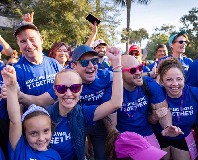 Group of smiling participants at an outdoor charity walk or run, wearing matching blue T-shirts with the slogan 'BUILDING HOPE TOGETHER.' The atmosphere is cheerful and energetic, with people cheering, some wearing sunglasses and hats, surrounded by trees and sunlight.