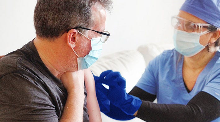 Person wearing scrubs, a surgical mask and gloves touches upper arm of man in gray t-shirt