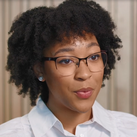 Black woman with curly shoulder-length hair, glasses, large blueish pearl earrings, white collared shirt 