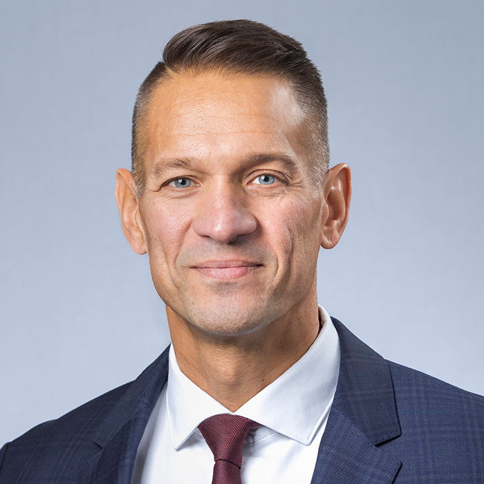 A headshot of a man in a suit, smiling slightly against a gray background