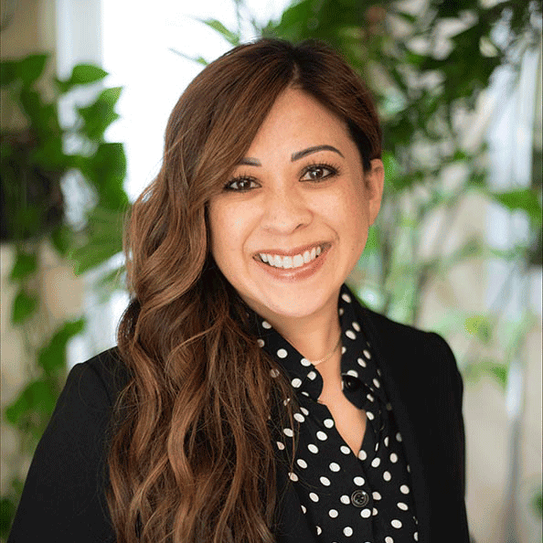 Headshot of a smiling woman with long brown hair, wearing a black blazer and polka dot blouse, with greenery in the background