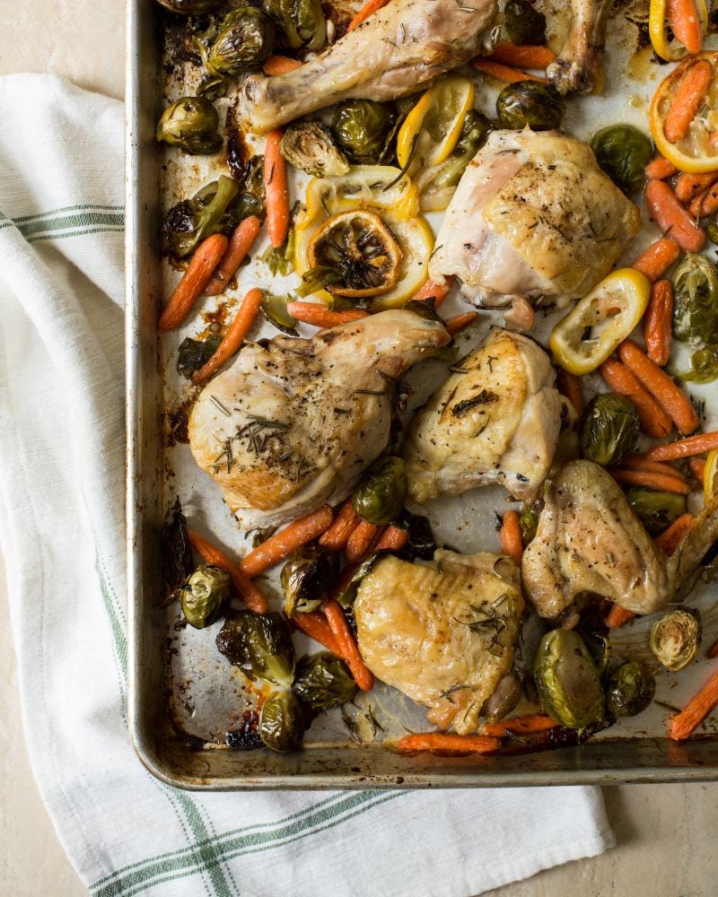Pieces of cooked chicken, baby carrots, Brussels sprouts, and sliced lemons fill a baking sheet that rests on a white dish towel.