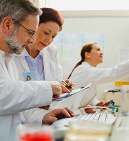 Dos investigadores examinan información juntos en un entorno de laboratorio y otro investigador está en el fondo de la imagen.