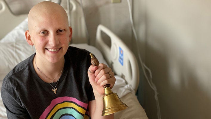 Paciente joven con cáncer sonriendo y sosteniendo una campana en una sala de hospital