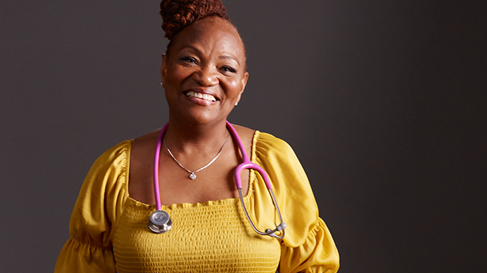  A woman in a yellow dress with a stethoscope around her neck, standing against a dark background