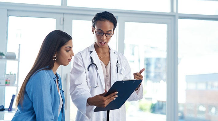 A woman with long hair looks at a clipboard held by a doctor standing in front of a wall of windows. 