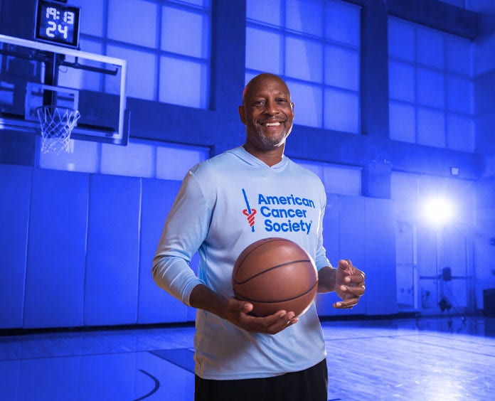 Alonzo Mourning, ex jugador de la Asociación Nacional de Baloncesto (NBA) y miembro del Salón de la Fama del Baloncesto, sostiene una pelota de baloncesto en la mano derecha con un jersey de la Sociedad Americana contra el Cáncer.