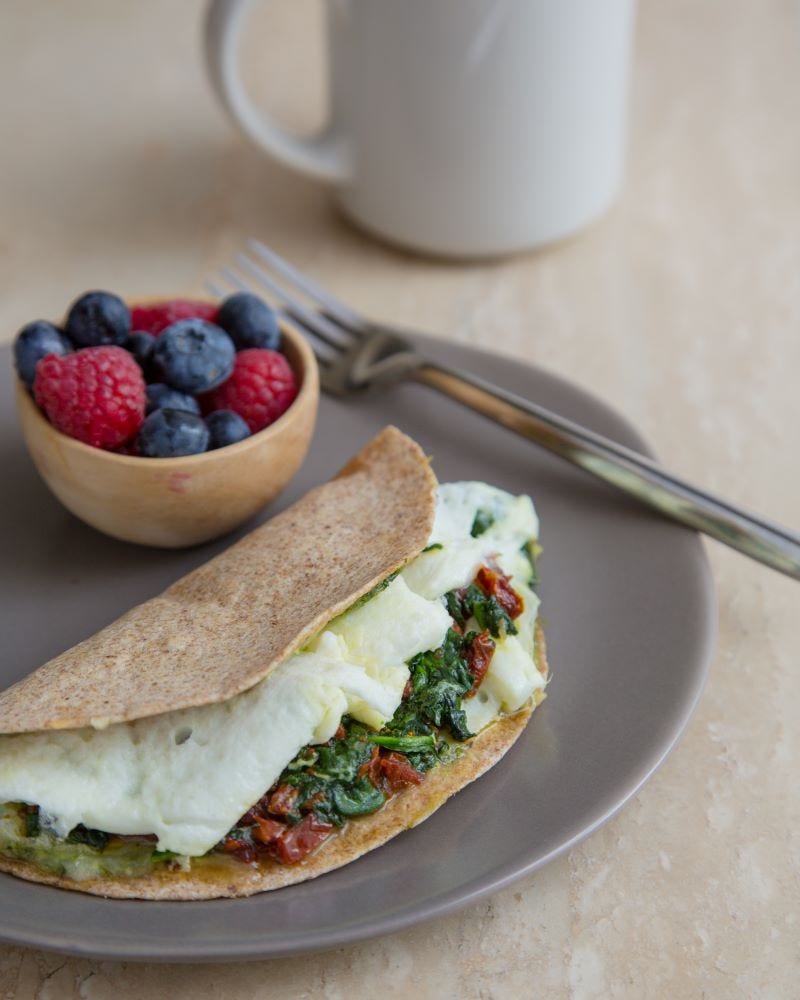 A whole wheat tortilla filled with egg whites, tomatoes, and spinach rests on a plate next to a fork and a small bowl of blueberries and raspberries.