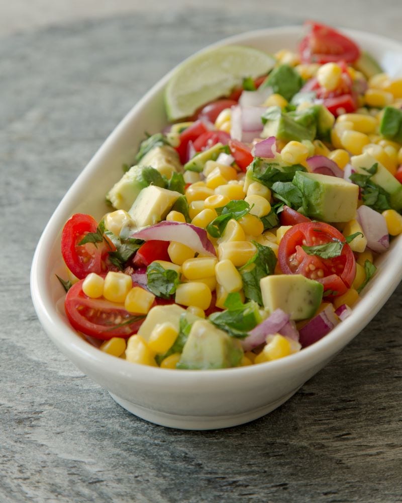 A mix of corn kernels, tomatoes, onions, avocados, and cilantro fills a white oblong bowl. 