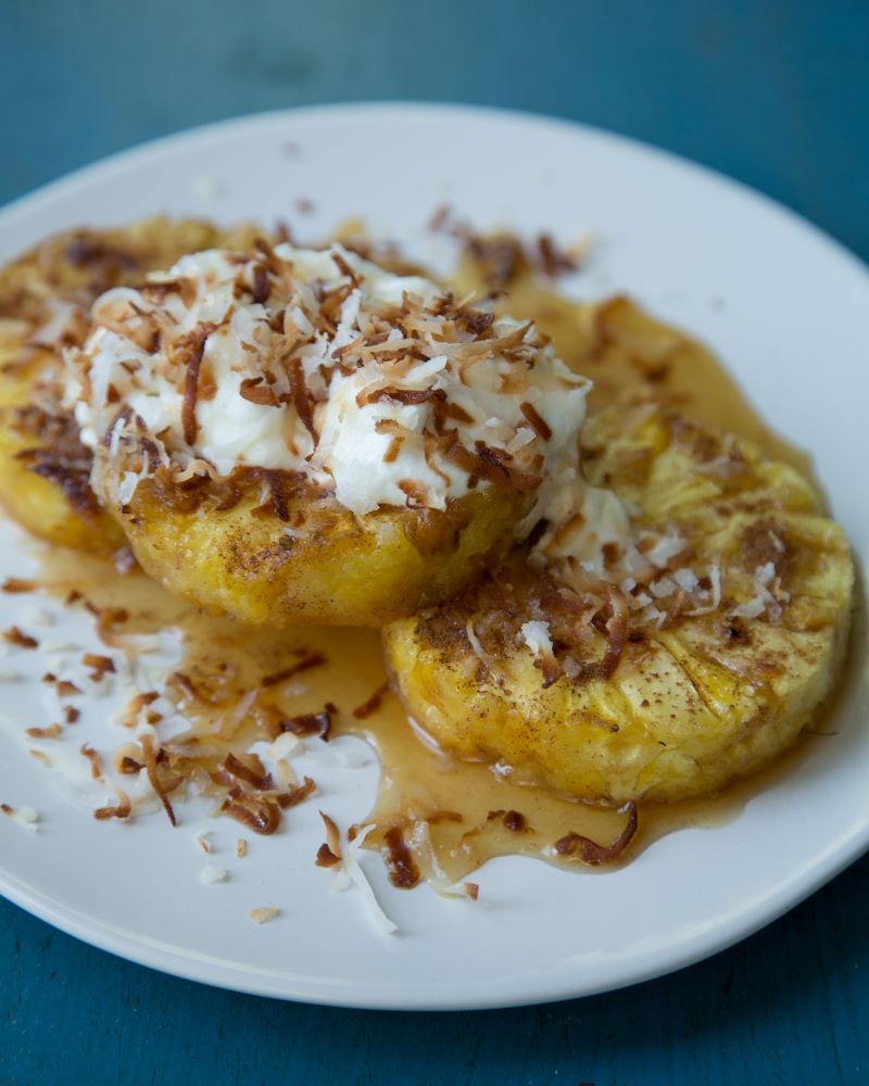 Slices of roasted pineapple topped with toasted coconut and plain yogurt sit on a round white plate.