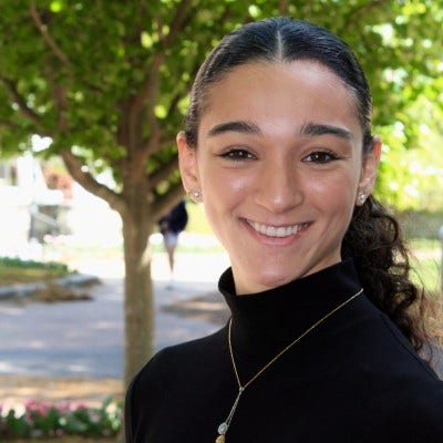 Woman in front of tree with hair parted in middle and pulled back in curly pony tail in black mock turtle neck long sleeved shirt and drop pearl necklace