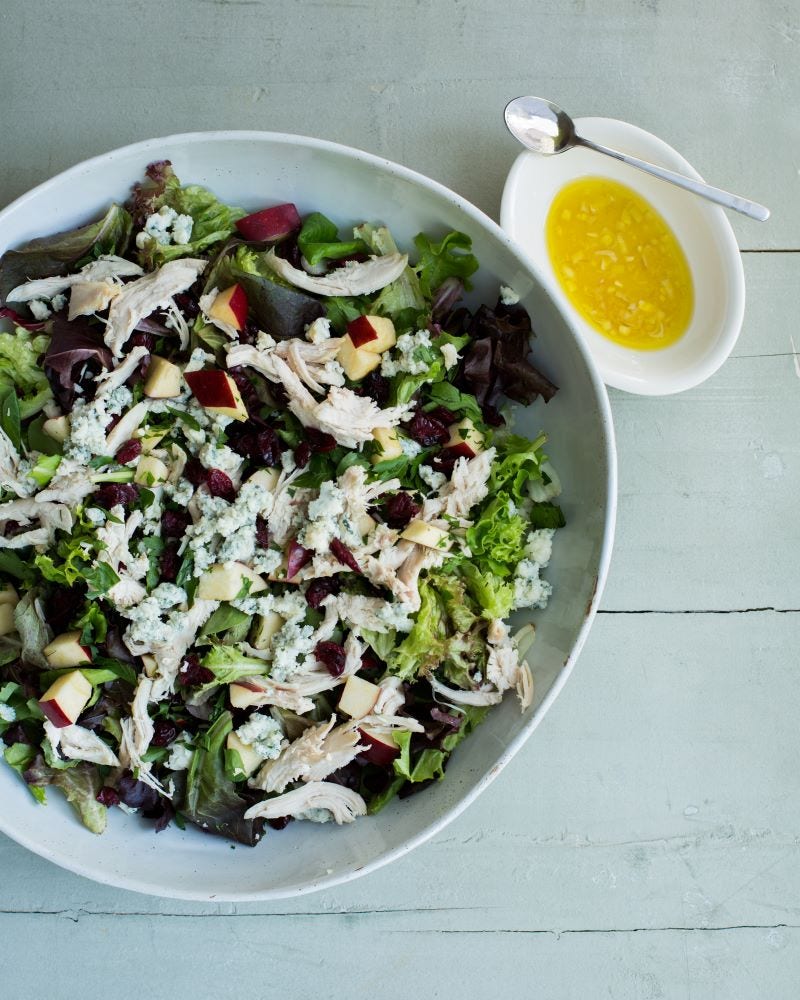 A salad of greens, shredded chicken, apple cubes, and blue cheese crumbles fills a salad bowl that rests next to a small dish of dressing on a table with wood slats. 