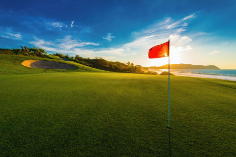 Golf flag on a well-maintained green at sunset, with a scenic ocean view and a sand bunker in the background