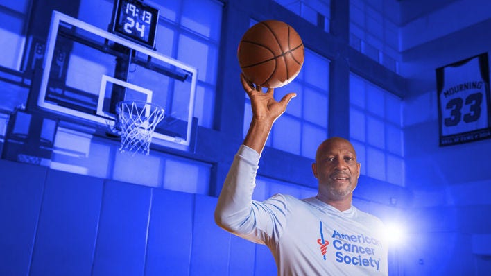 Alonzo Mourning, ex jugador de la Asociación Nacional de Baloncesto (NBA) y miembro del Salón de la Fama del Baloncesto, sostiene una pelota de baloncesto en la mano derecha con un jersey de la Sociedad Americana contra el Cáncer.