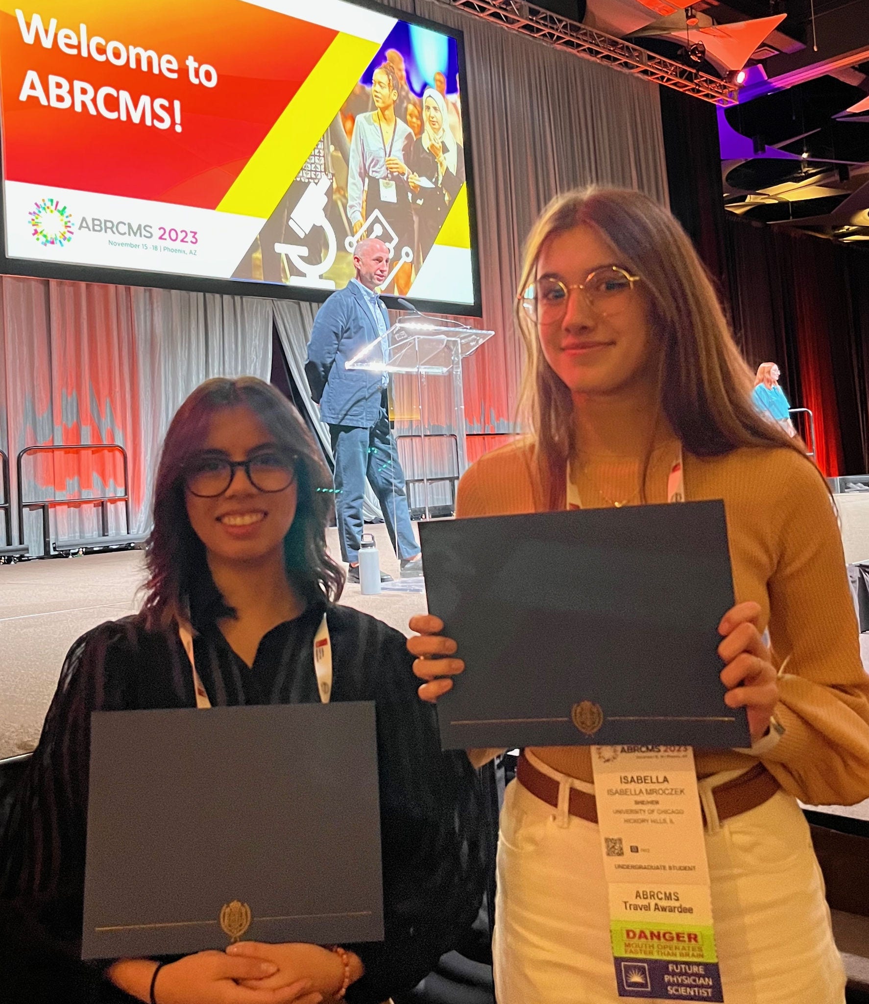 2 women one tall holding cray folders in front of stage with Welcome to ABRCMS on screen