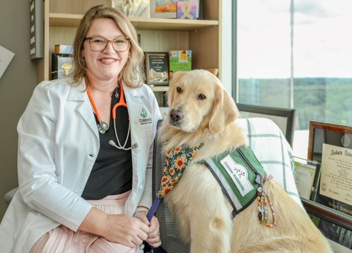 Smiling doctor and golden retriever sit in an office with children's books on shelves behind them.