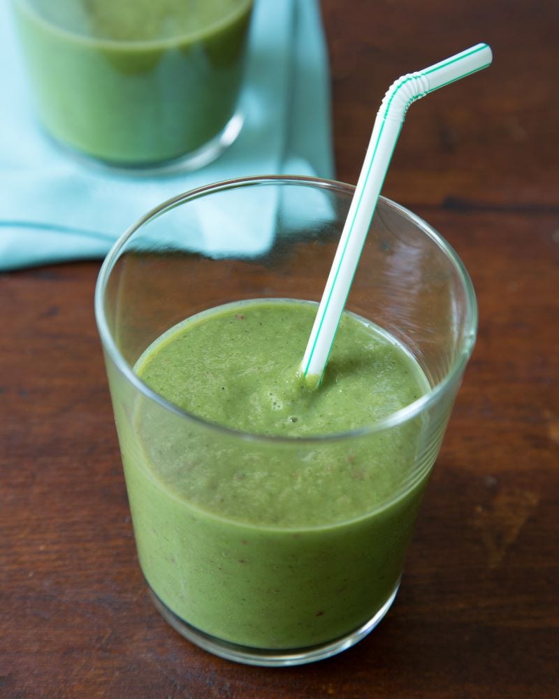 A bent straw sticks out of a green shake in a glass that sits in front of another green shake on a table.
