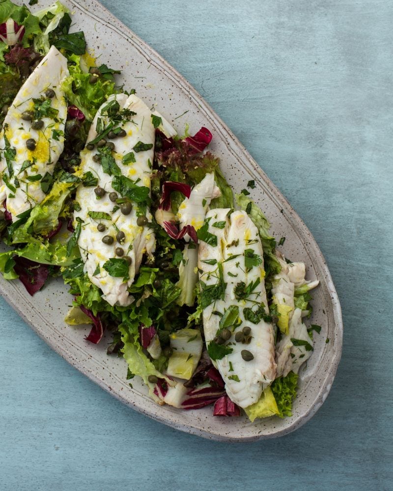 Fish filets topped with herbs lay on a bed of lettuce on an oval-shaped earthenware platter.