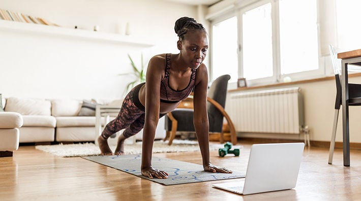 A woman does a pushup while looking at a laptop computer on the floor of a living room. 