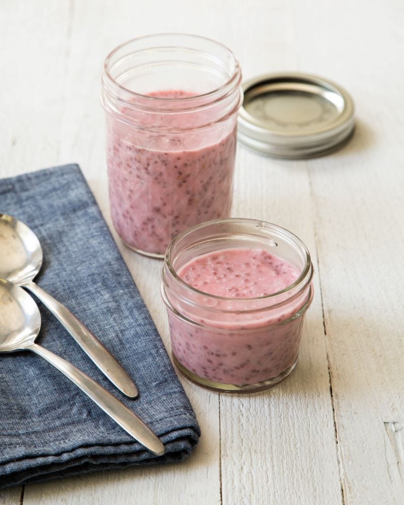 Two small jars of raspberry chia pudding sit on a rustic wood table next to a folded blue napkin and two spoons. 