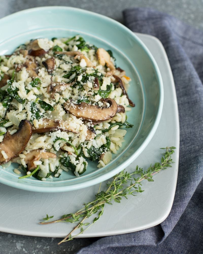 A bowl of spinach, mushrooms, risotto, and Parmesan cheese sits on a square plate next to several sprigs of thyme. 