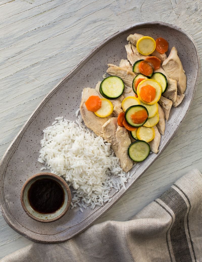 Strips of steamed chicken breast topped with slices of squash, zucchini, and carrots lay next to white rice and a cup of sauce on a platter. 