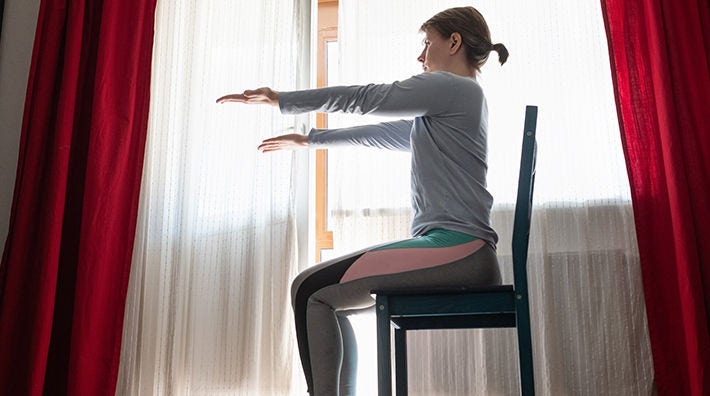 A woman extends her arms while sitting in a chair in front of a window with sheer curtains. 