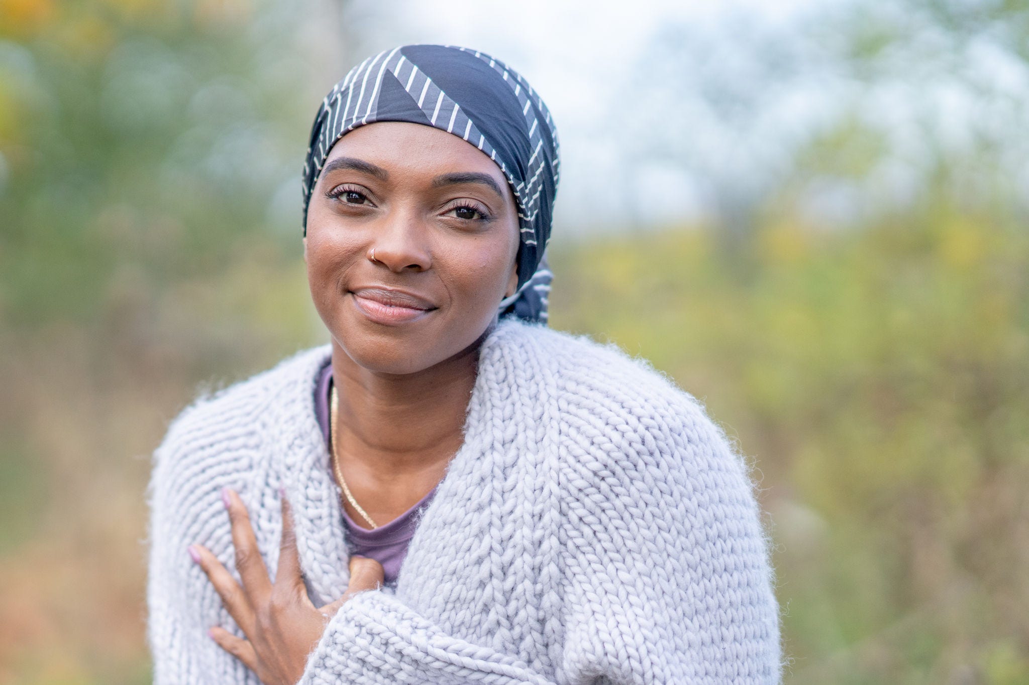 A young woman of African decent, who is battling cancer, sits outside on a cool fall day.  She is dressed warmly in a sweater and head scarf as she enjoys the fresh air between treatments.