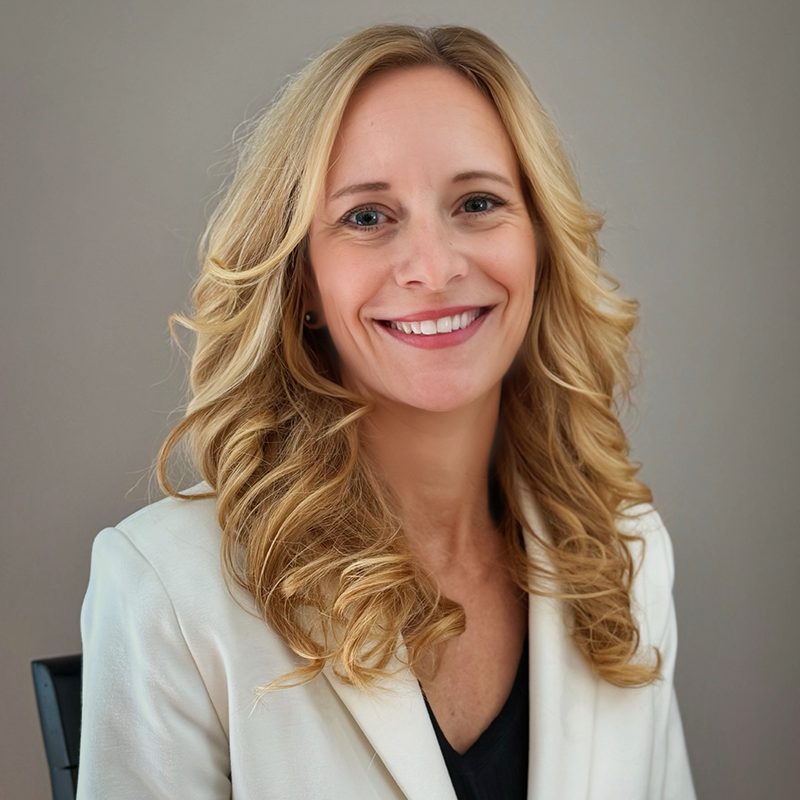 A woman with blonde hair smiles while wearing a light-colored blazer.