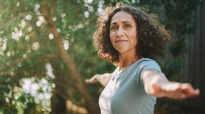 Woman in gray t-shirt extends arms while standing in front of trees 