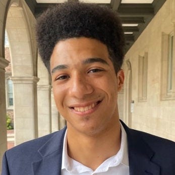 black young man in gray jacket over white shirt next to columns of building