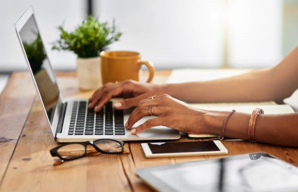 Stock image of a person's hands working on a laptop, black frame eyeglasses and smartphone in the foreground