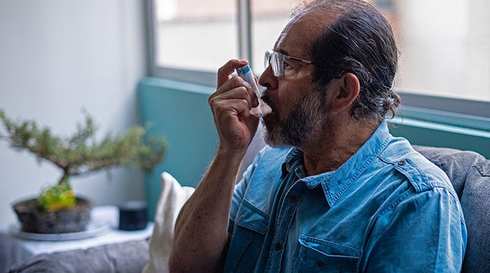 An older man with a beard and glasses uses an asthma inhaler as he sits on a couch 