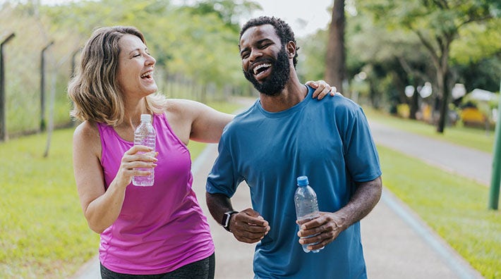 A woman and a man in workout clothes hold water bottles and laugh as they walk through a park. 