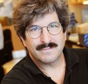 white man with round wire rim glasses, moustache, and v-neck collared brown shirt