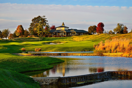 A golf course featuring a clubhouse on a hill, surrounded by autumn foliage, with a water feature in the foreground