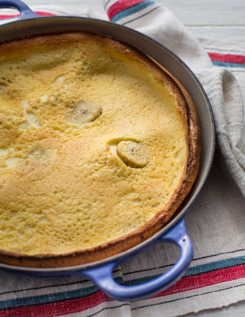 A banana Dutch baby fills a round cooking pan with blue handles that rests on a woven cloth with striped edges. 