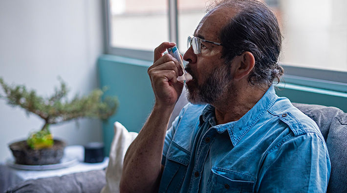 An older man with a beard and glasses uses an asthma inhaler as he sits on a couch 