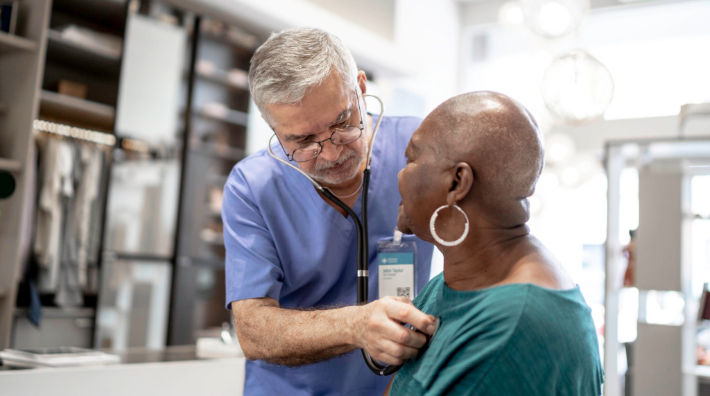 A male doctor wearing blue scrubs listens the heart of a woman wearing a green T-shirt.