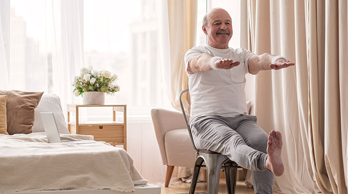 Bald man with moustache sits in a chair and extends arms and one leg