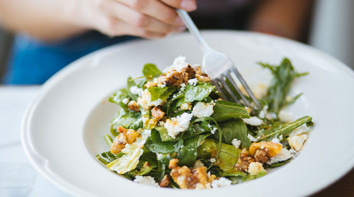 A woman’s hand holds a fork in a green salad in a white bowl