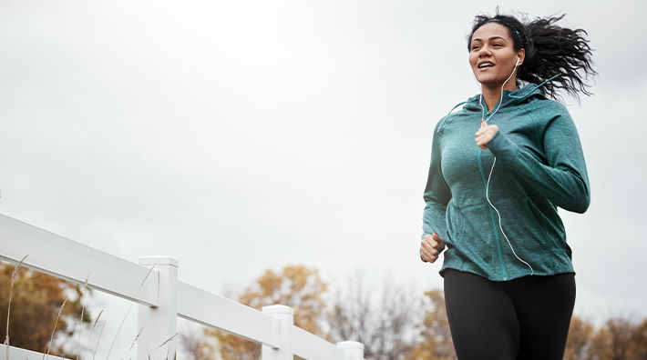 A woman wearing a green jacket and listening to headphones jogs next to a white fence 