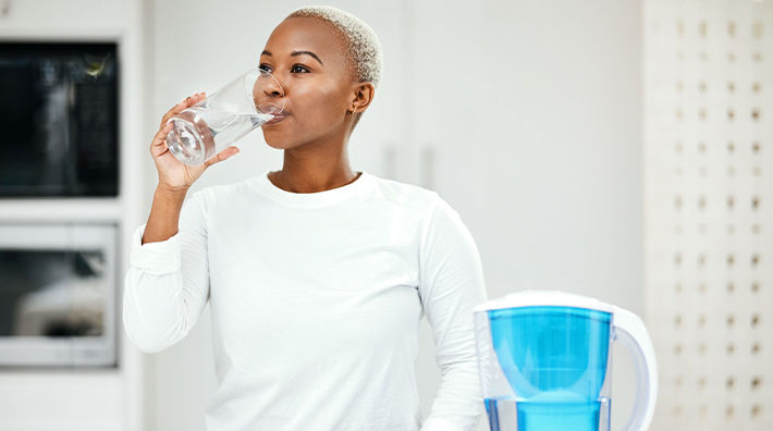 A woman wearing a white shirt drinks a glass of water from a pitcher next to her.