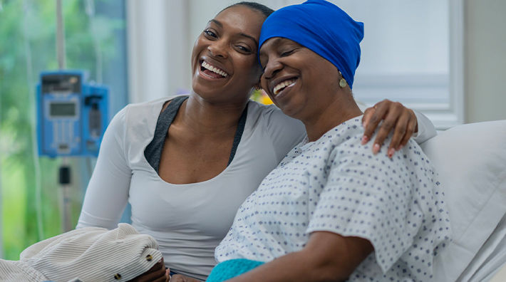 A smiling woman has her arm around an older woman wearing a headscarf and a medical gown in a hospital bed 