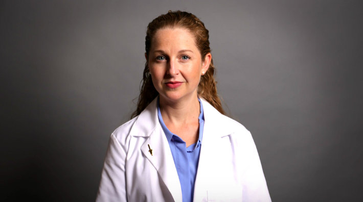 Cancer researcher Dr. Charly Good wears a lab coat and stands in front of a gray background