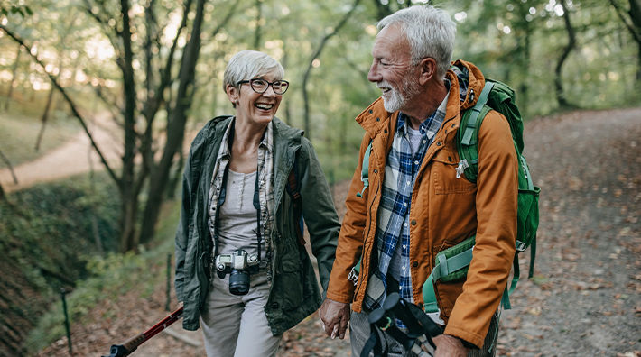 An older man and woman hold hands and smile at each other while walking on a nature trail. 